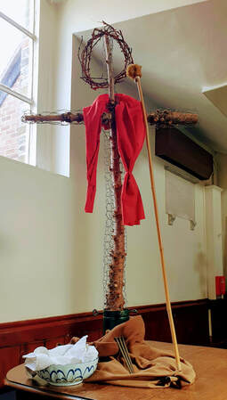 A cross made from the trunk of a Christmas tree, dressed with lenten symbols including a crown of thorns, a bowl and towel, nails, and a stick with sponge on the end.